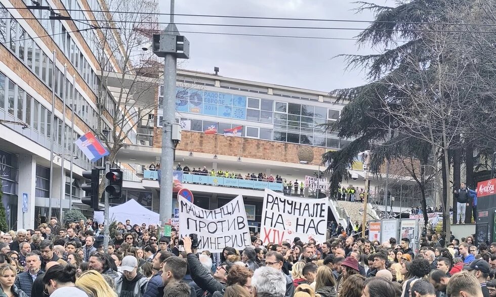Photo: Protest in Belgrade. A large crowd holds signs with Serbian text. Security forces stand on a staircase in front of a building.